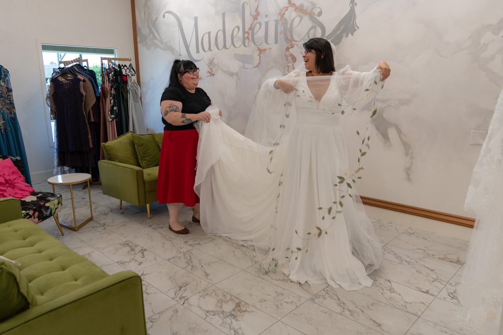 bridal store employee helping bride try on wedding dresses and veils